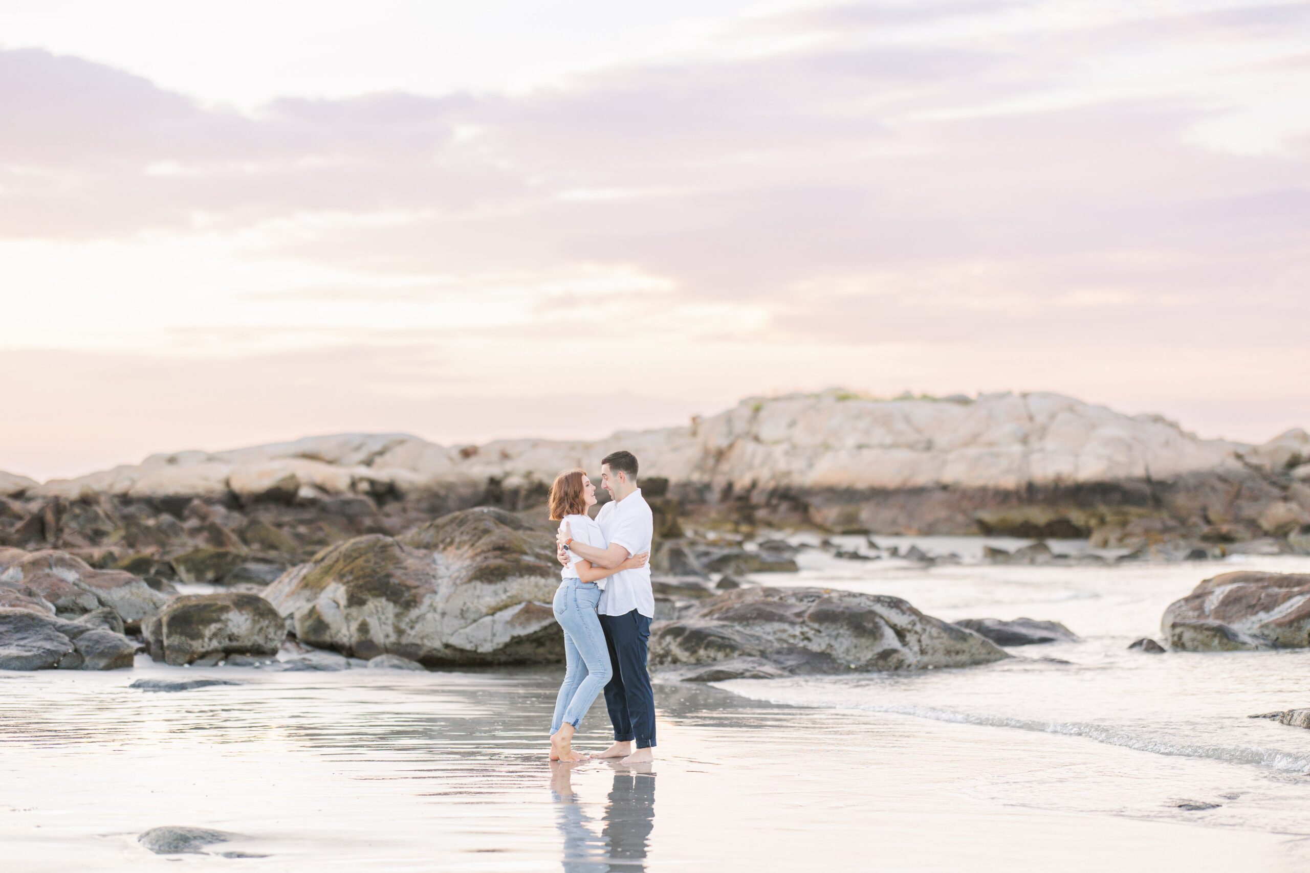 A couple hugs while standing in shallow water at Wingaersheek beach, surrounded by large rocks and a sunset sky.