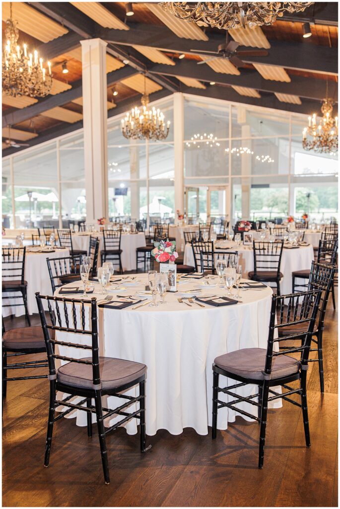 Indoor wedding reception with round tables, white linens, and black chairs under chandeliers.