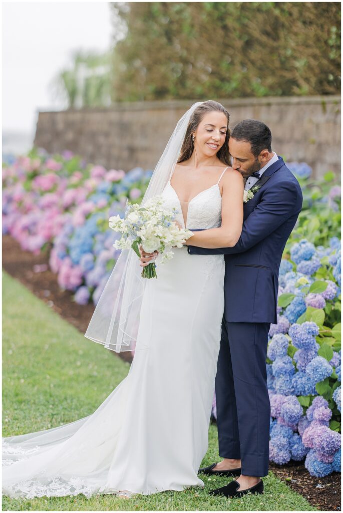 Groom hugging bride from behind in front of colorful hydrangeas at their wedding.