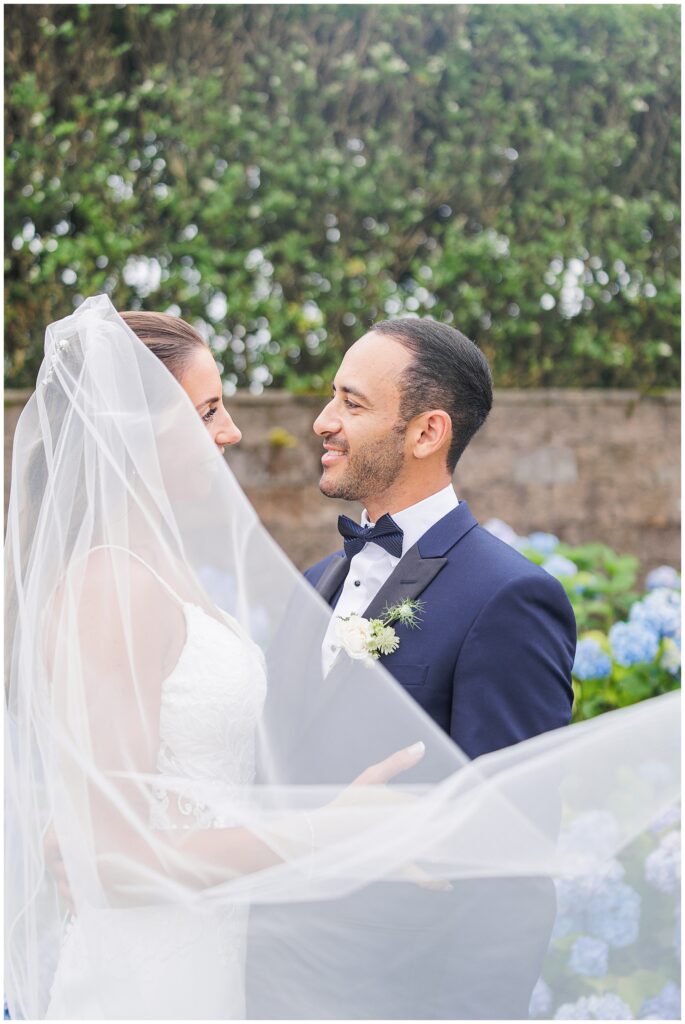 Bride and groom smiling at each other, bride’s veil draped over them.