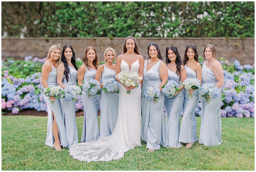 Bride with her bridesmaids holding blue hydrangea bouquets in front of hedges at the Wychmere Beach Club.