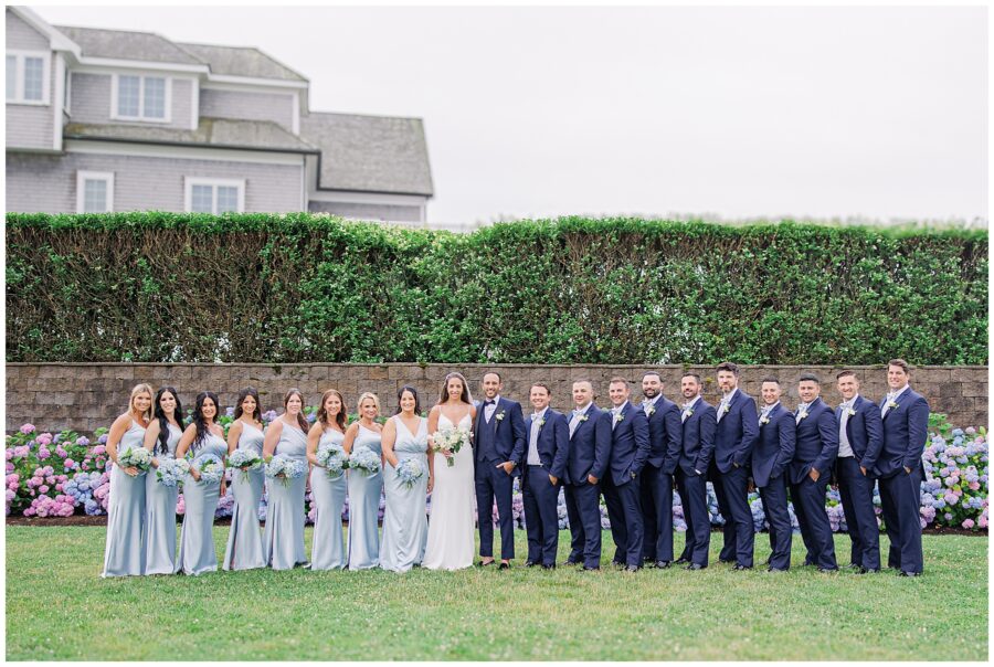 Full wedding party standing in front of hedges with hydrangeas.