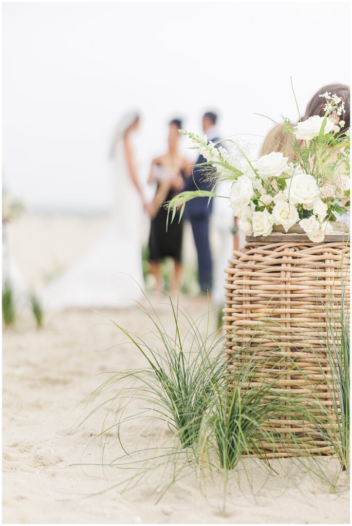 Wedding ceremony on the beach, focus on floral arrangement in the foreground.