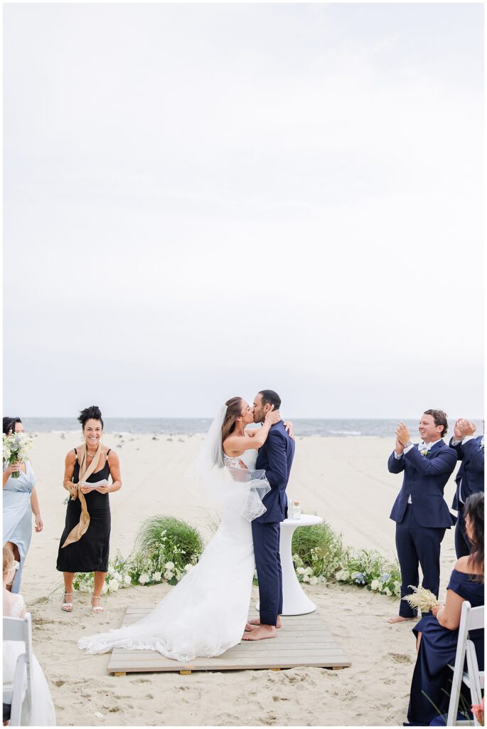 Bride and groom kiss on a beach platform as guests clap at Wychmere Beach Club.