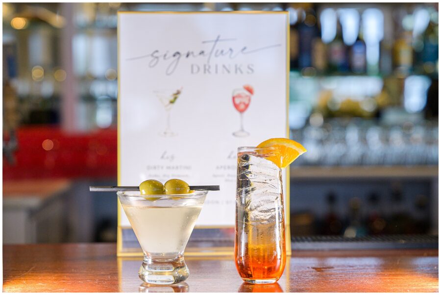 Two signature cocktails on a bar with a sign in the background: a martini with olives and an orange cocktail.