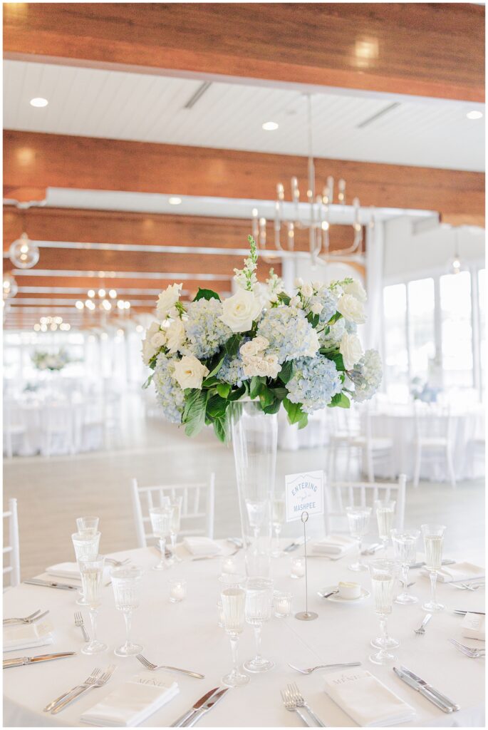 Table setting with large floral centerpiece of white roses and blue hydrangeas at a wedding.