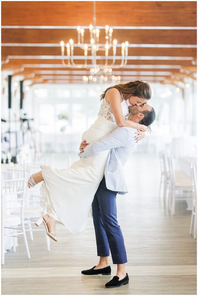 Groom lifts bride for a kiss in the Harbor Room with chandeliers and white chairs at a Wychmere Beach Club wedding.