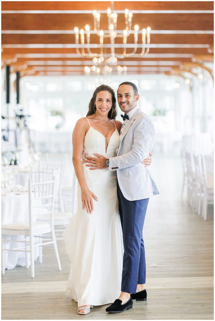 Bride and groom embrace in elegant reception hall with chandelier overhead. Beach club wedding venue on Cape Cod.