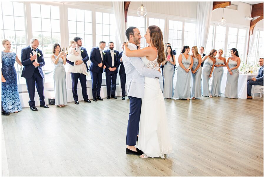 Newlyweds share first dance surrounded by wedding guests. Intimate moment in bright reception space.