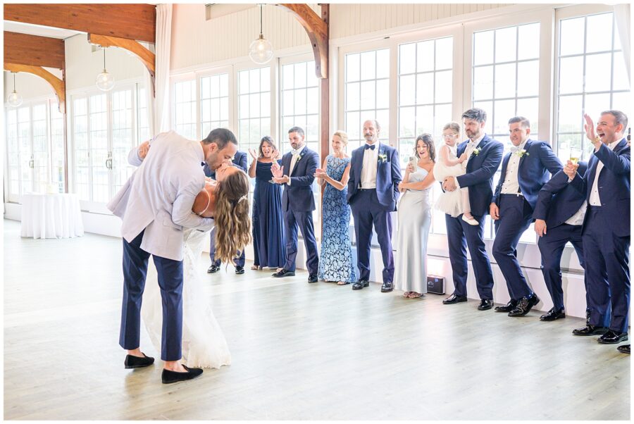 Wedding couple kissing while guests cheer. Elegant venue with large windows and wooden beams.