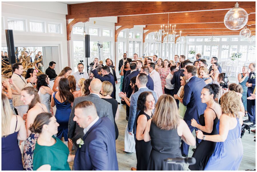 Lively wedding reception with many guests dancing on the dance floor. Wooden beams and chandeliers visible.