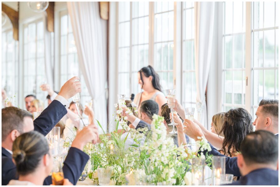 Wedding guests raising glasses for a toast during reception dinner. Floral centerpieces on tables.