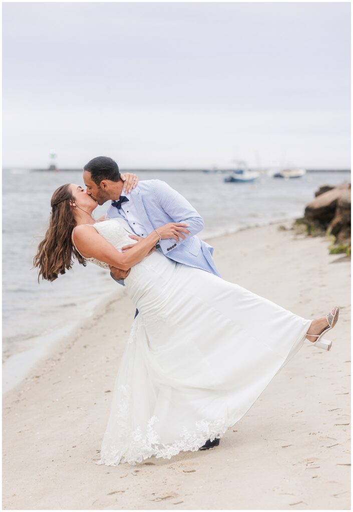 Bride and groom kissing on beach, groom dips bride dramatically. Ocean and boats visible in background.