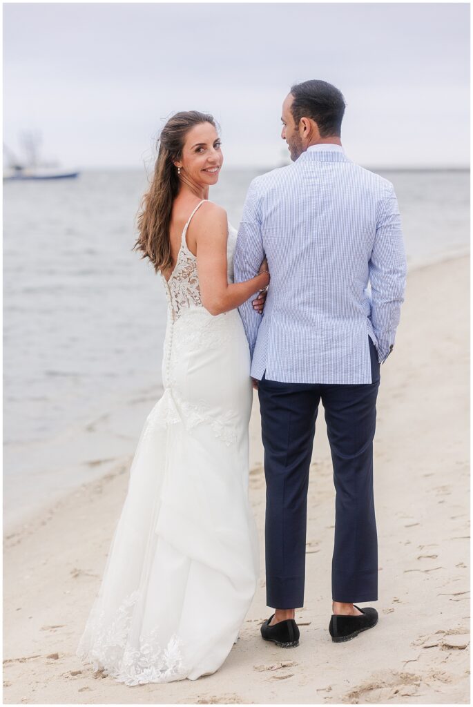 Bride and groom walking hand-in-hand on beach, bride smiling at camera. Coastal setting with water visible.