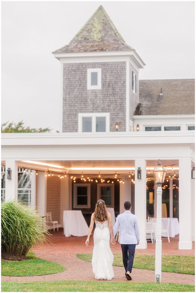 Couple walking towards Wychmere Beach Club Harbor Room. String lights and Cape Cod architecture visible. Evening wedding scene.