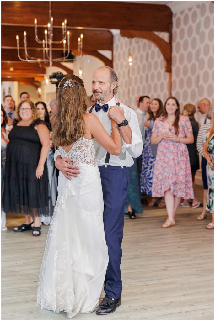 Bride and father dancing at wedding reception. Wooden beams, chandelier, and guests in background.