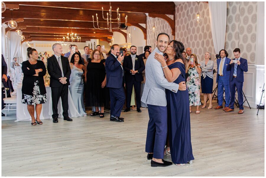 Couple dancing at wedding. Guests watching and taking photos. Elegant venue with chandeliers.