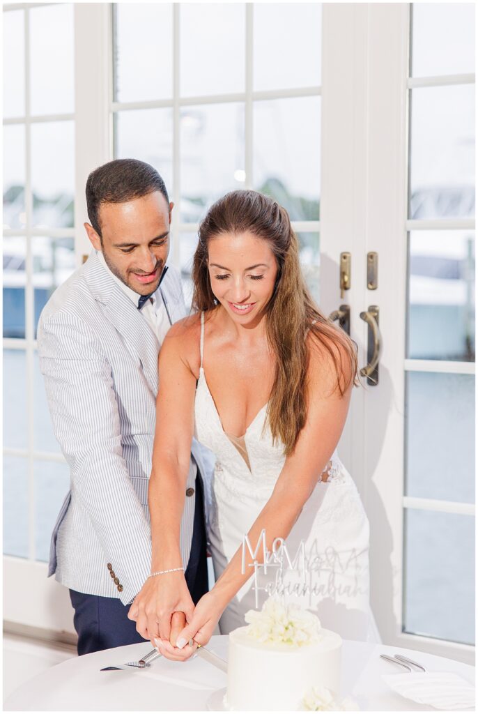 Newlyweds cutting cake together at Wychmere Beach Club. Large windows overlooking marina in background.