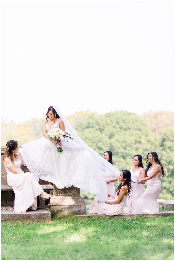 Bride sits on a stone ledge holding a bouquet as five bridesmaids in pale pink dresses adjust her flowing gown outdoors.