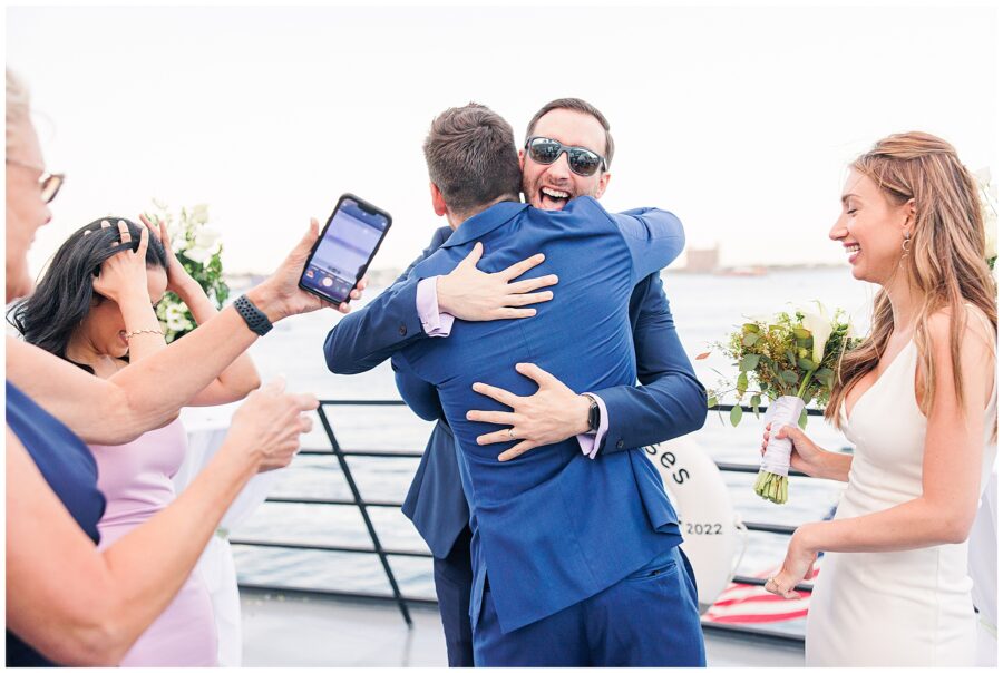 Two men in suits hug on a boat as others smile and take photos, with a waterfront view in the background.