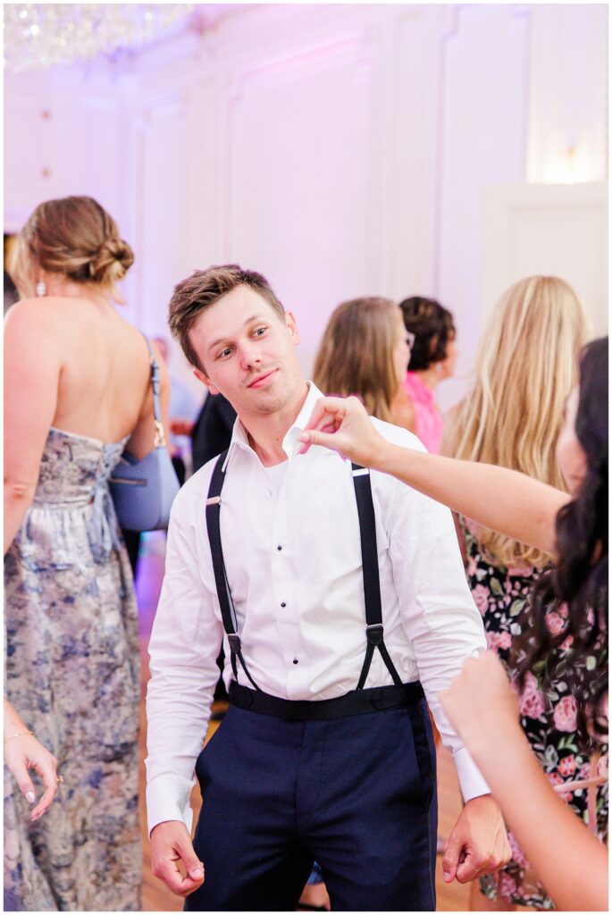 A man in a white shirt and suspenders dances at a wedding reception, surrounded by other guests in formal attire.