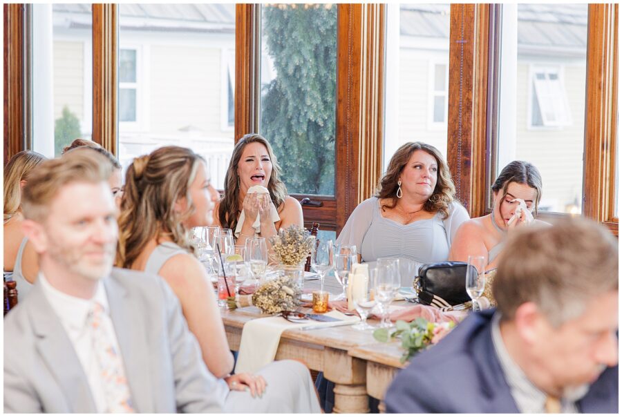 Guests seated at a wooden table react emotionally during a wedding reception, some wiping away tears.