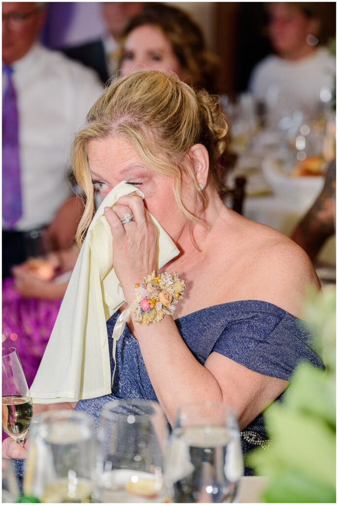 A woman wipes tears with a napkin during a wedding reception, holding a glass of champagne in the other hand.