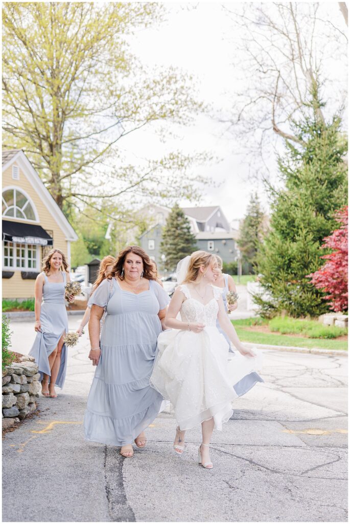 Bride in a white gown walks with bridesmaids in pale blue dresses on a tree-lined street, holding her dress up slightly.