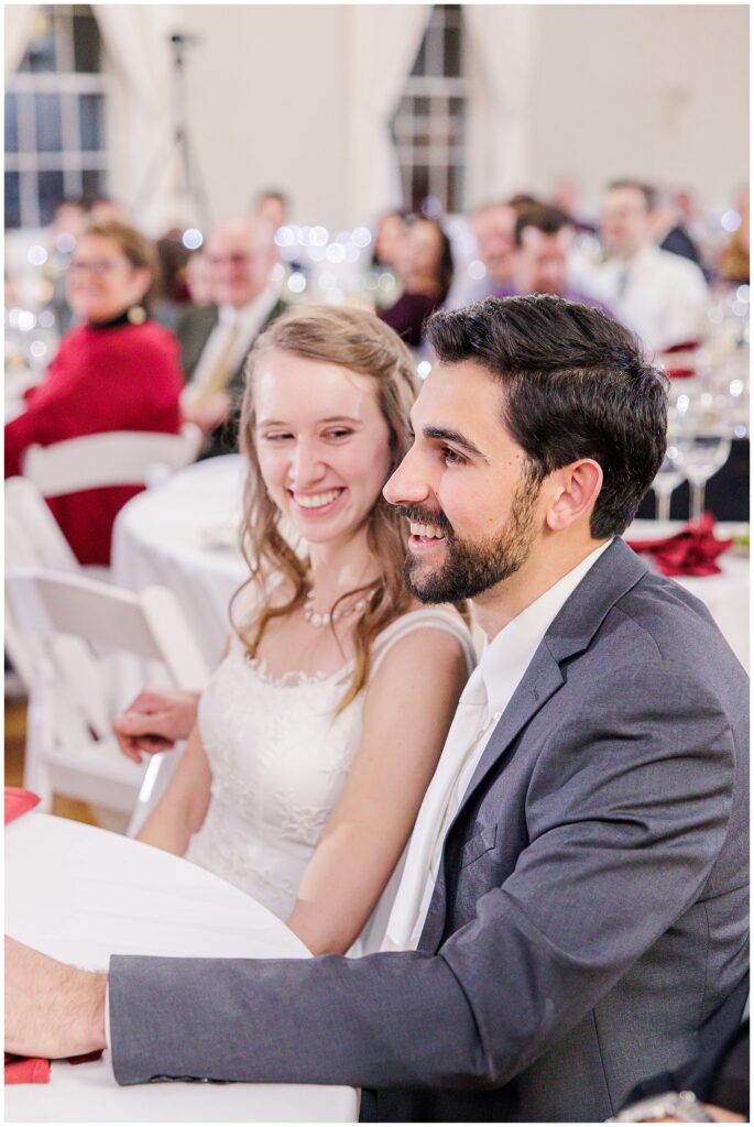 Bride and groom smile while seated at a reception, surrounded by guests at tables with white tablecloths and decor.