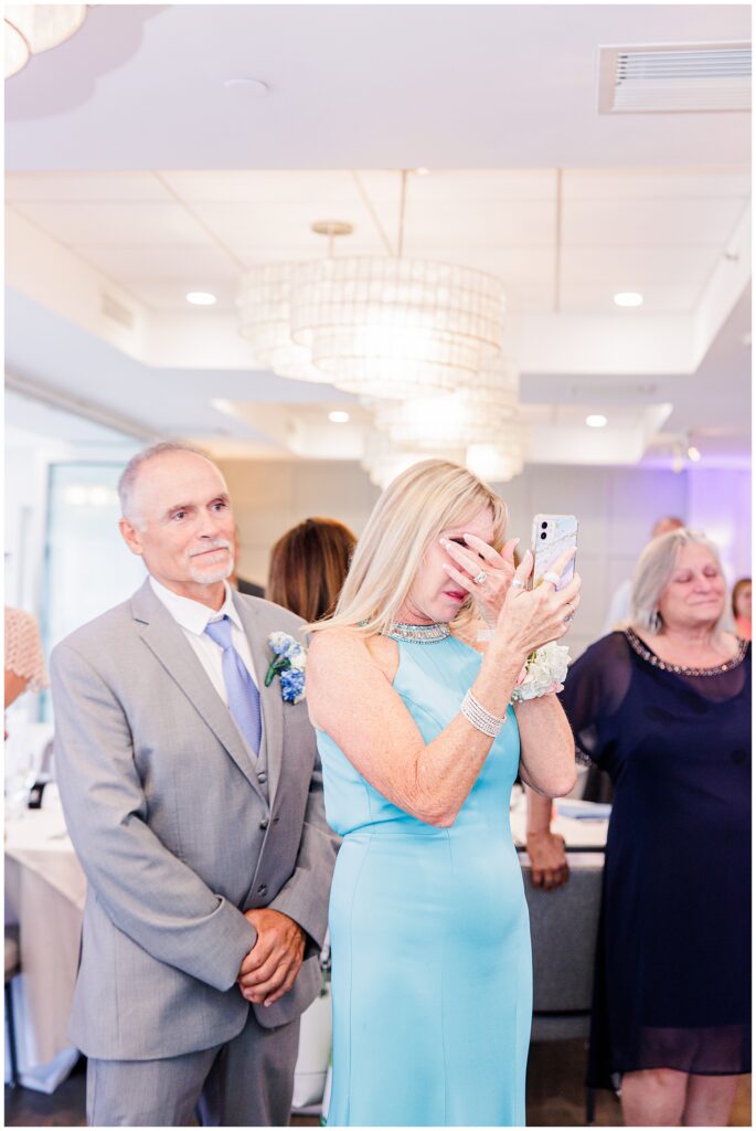 Woman in a light blue dress wipes tears while taking a photo on her phone during a wedding, standing next to a man in a suit.