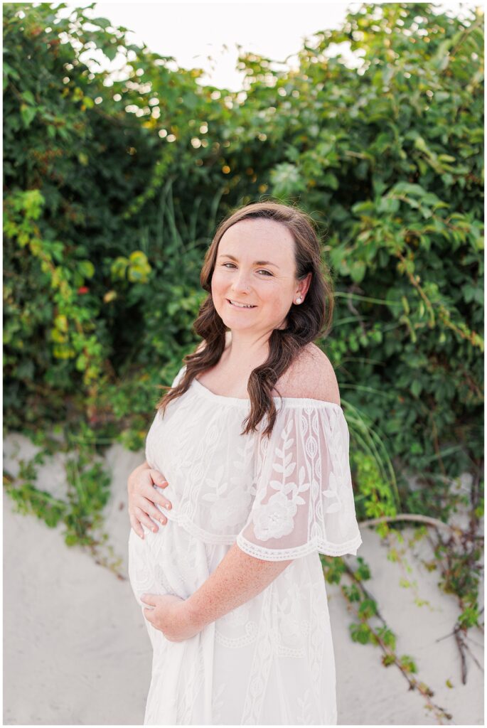 Pregnant woman in a lace dress holding her belly, smiling gently, with greenery in the background.