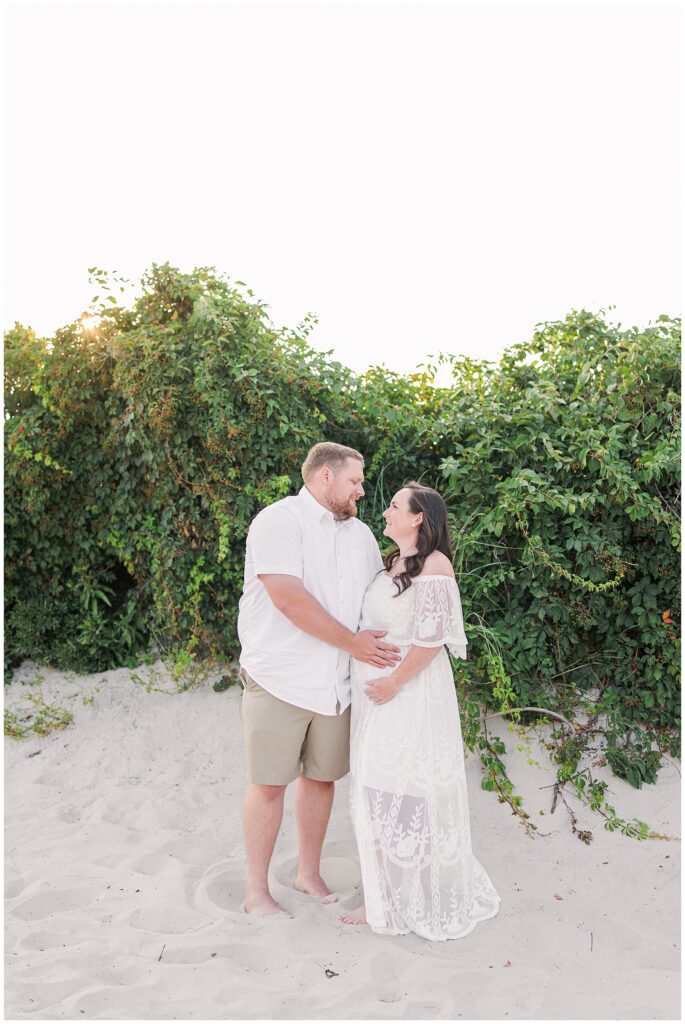 Couple smiling at each other, woman holding her pregnant belly, standing barefoot on sandy beach.