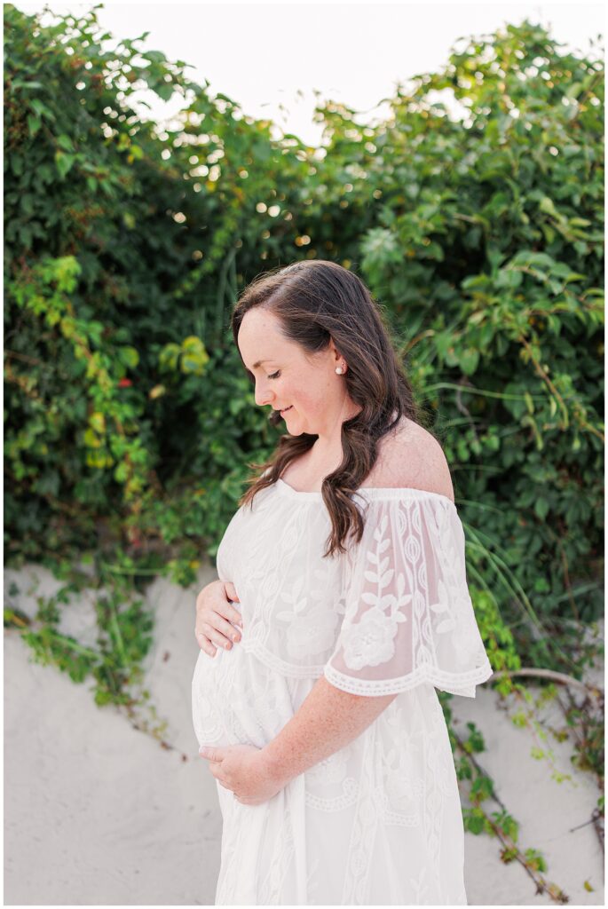 Close-up of a pregnant woman in a lace dress holding her belly, looking down, surrounded by greenery.