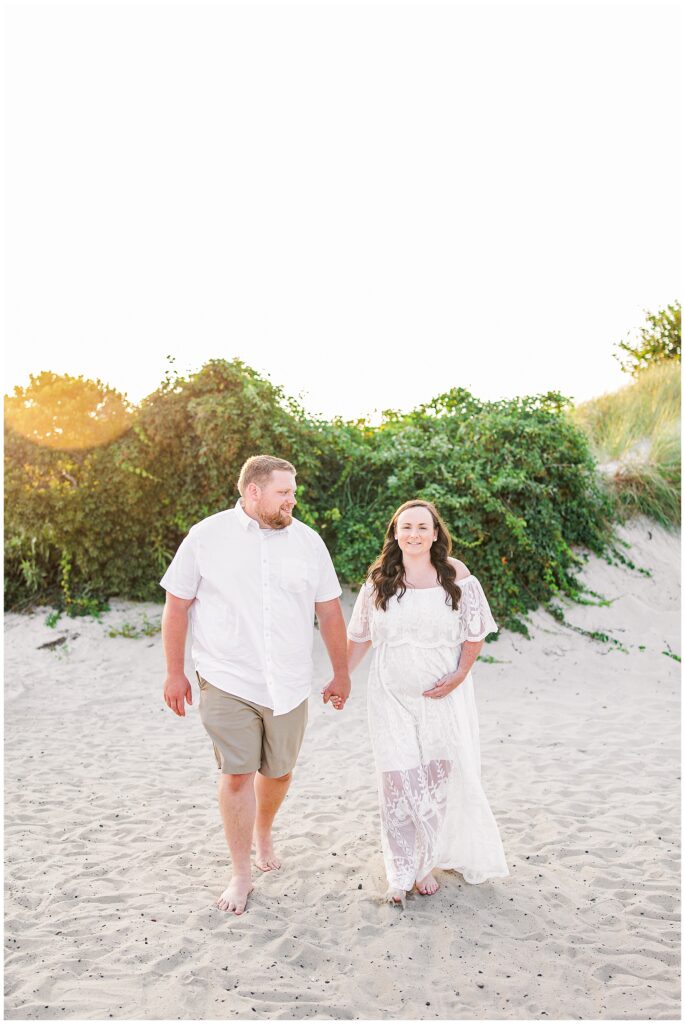 Couple walking hand in hand on the beach, woman holding her pregnant belly, sunlight in the background.