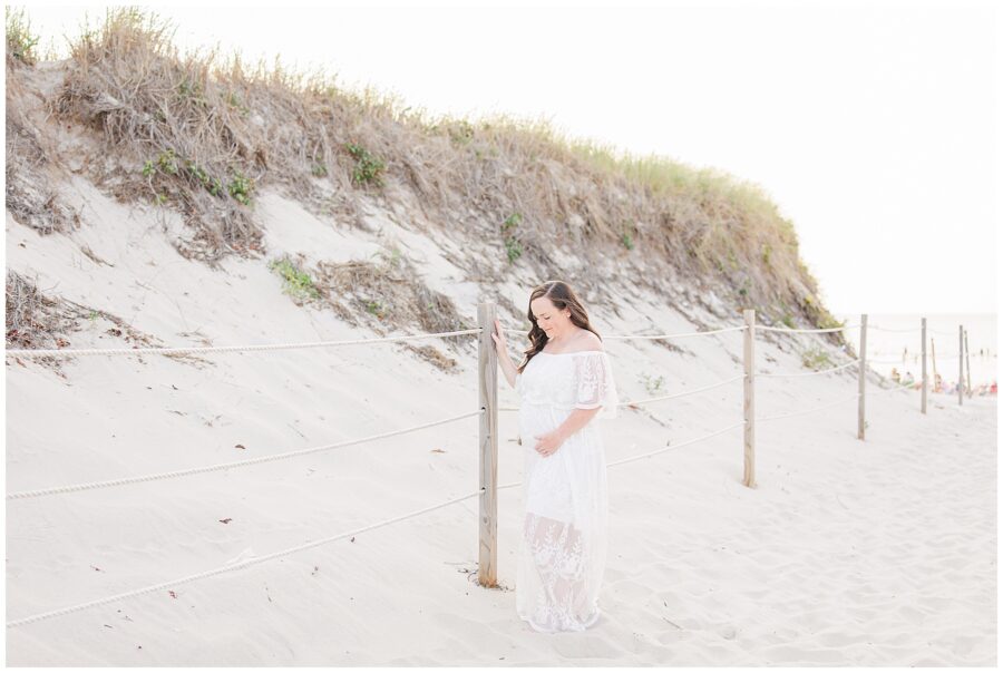 Pregnant woman in a lace dress standing by sand dunes, hand resting on a wooden post.