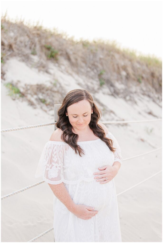 Pregnant woman in a lace dress standing by dunes, looking down, gently holding her belly.