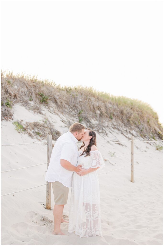 Couple kissing on a sandy beach, woman holding her pregnant belly, surrounded by sand dunes.