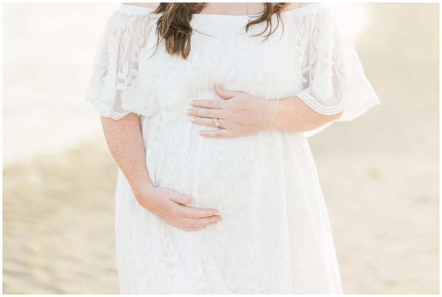 Close-up of a pregnant woman’s belly, hands gently resting on it, wearing a lace dress at the beach.