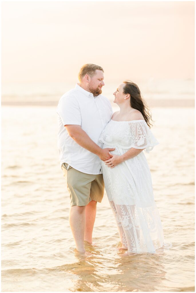 Couple standing in shallow water, smiling at each other, the woman holding her pregnant belly.