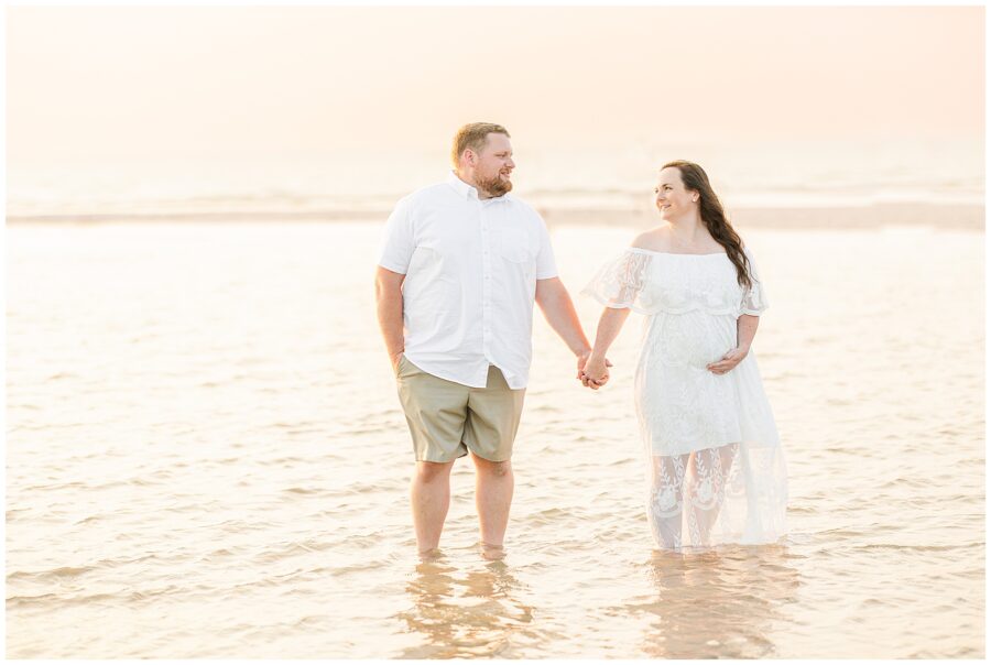 Couple holding hands, standing in shallow water at sunset, the woman cradling her pregnant belly.