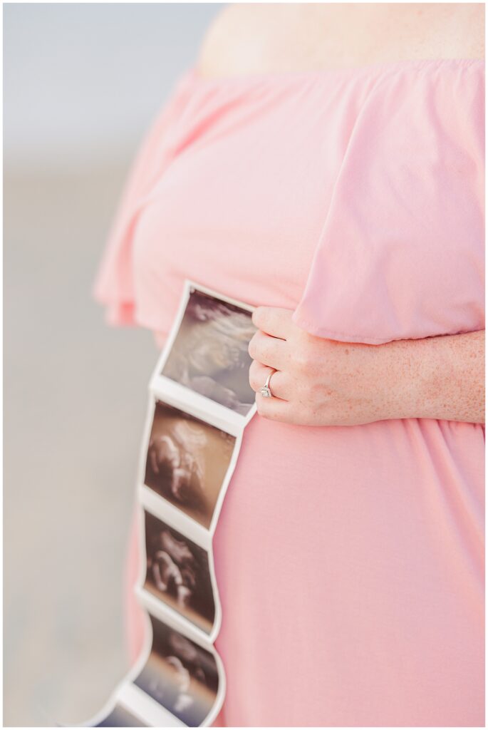 Close-up of a pregnant woman in a pink dress holding an ultrasound photo against her belly.