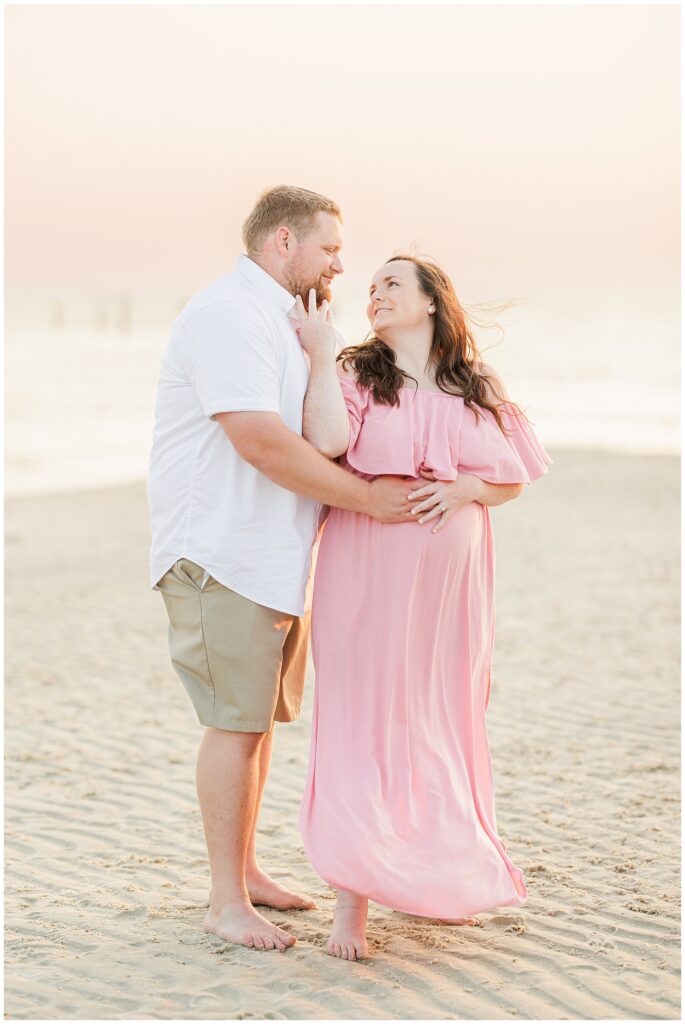 Couple standing on the beach at sunset, the woman in a pink dress, smiling and cradling her belly.