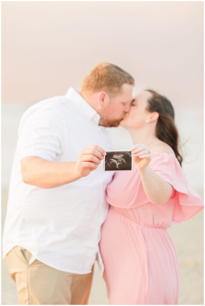 Couple kissing on the beach, holding an ultrasound photo, the woman wearing a pink dress.