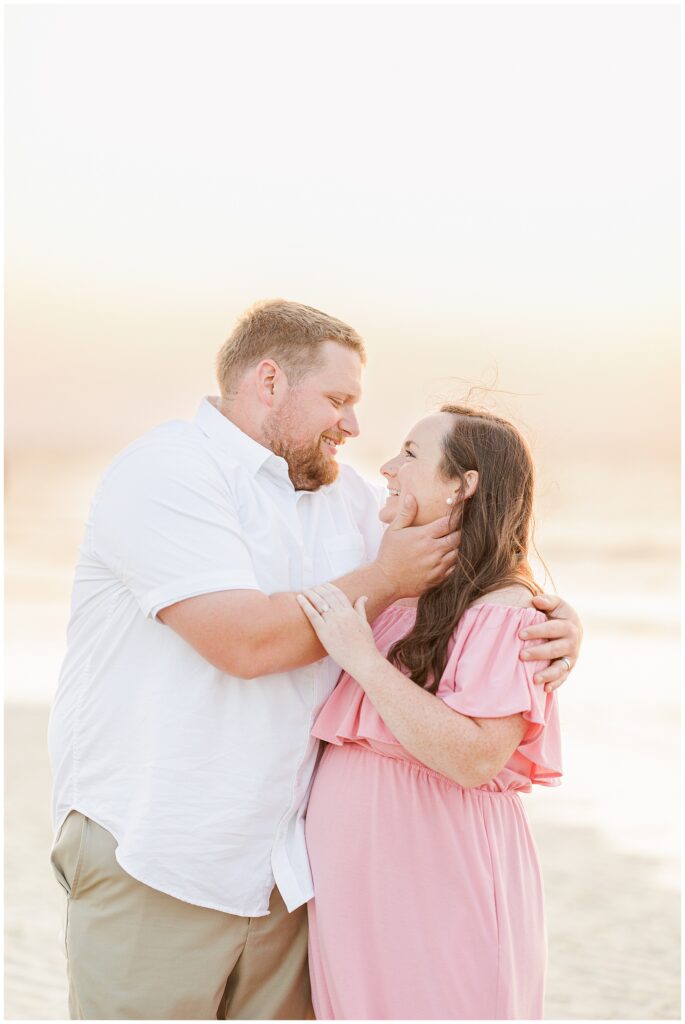 Couple embracing on the beach, smiling at each other, the woman in a pink dress, cradling her belly.