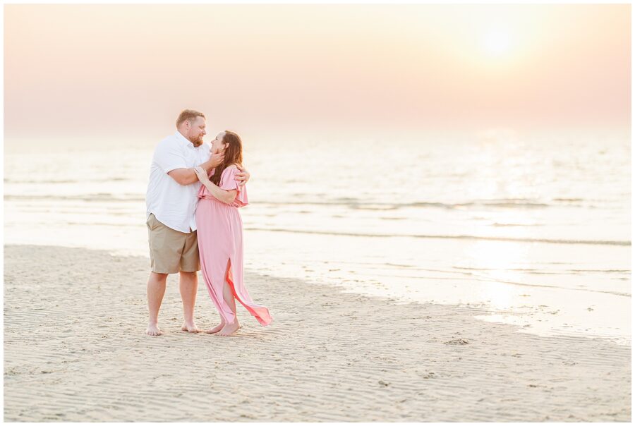 Wide shot of a couple embracing on the beach at sunset, with the woman in a flowing pink dress.