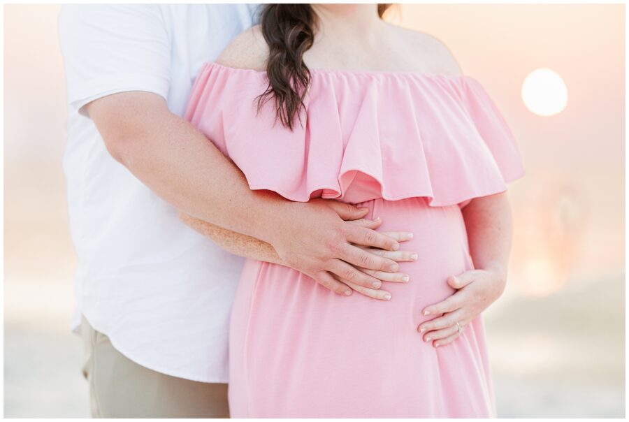 Close-up of a couple’s hands resting on the woman’s pregnant belly, both dressed in light colors at sunset.