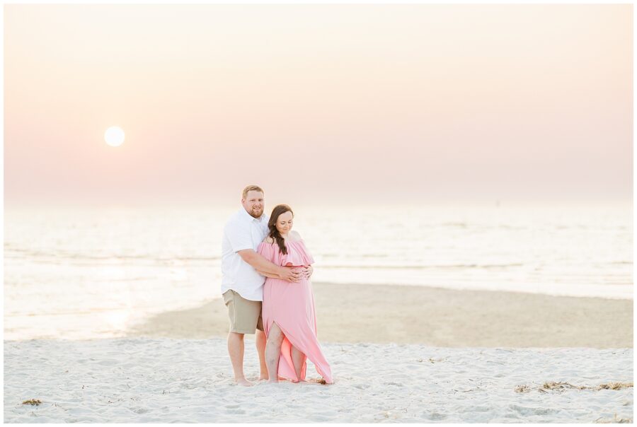 Cape Cod maternity photography of a couple on the beach at sunset, the woman in a pink dress, with the man holding her belly.