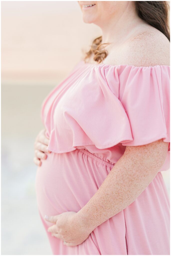 Close-up of a pregnant woman in a pink dress gently holding her belly, smiling.