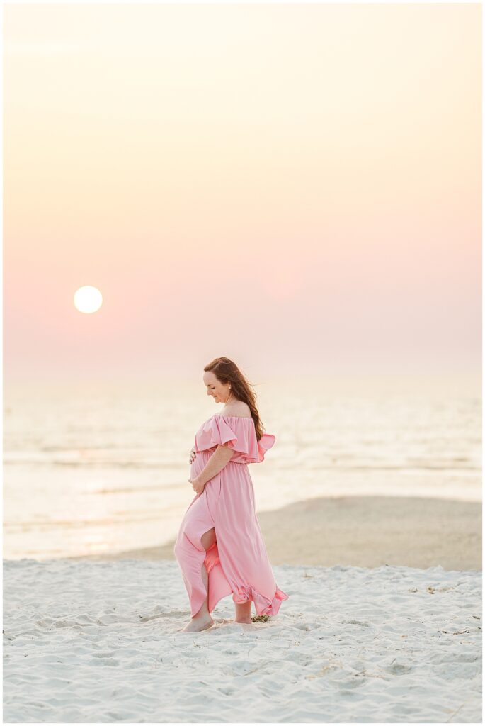 Pregnant woman in a flowing pink dress walking barefoot on the beach at sunset, cradling her belly.