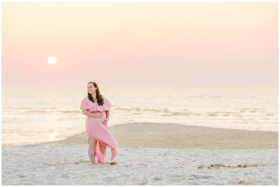 Woman in a pink dress standing on the beach at sunset, holding her belly, with a serene expression.